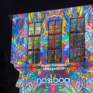 The Nosiboo logo displayed with colourful light on a building, informing about the bronze level of sponsorship of the Zsolnay Light Festival in Pécs.